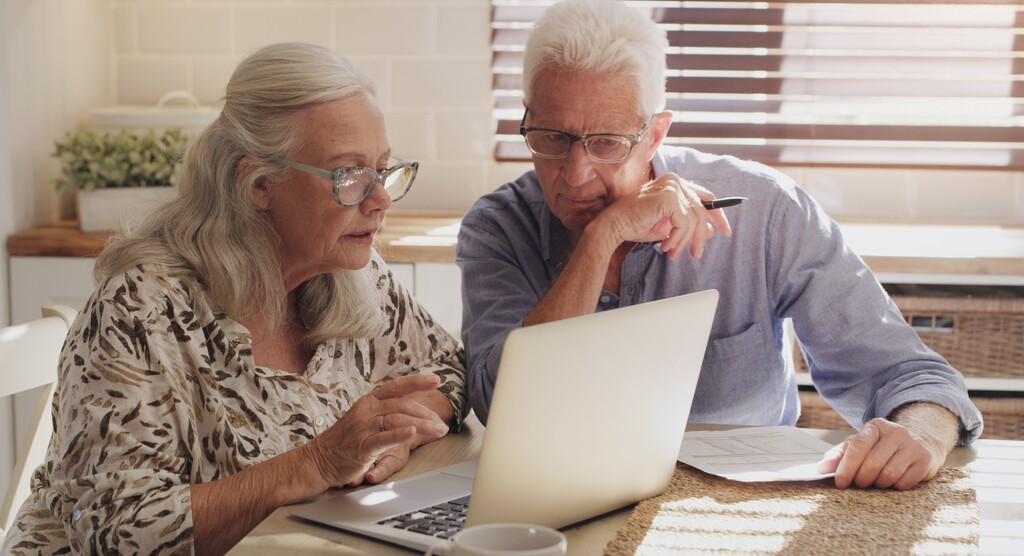 A man and a woman sit together at a table, reading information on a shared laptop.