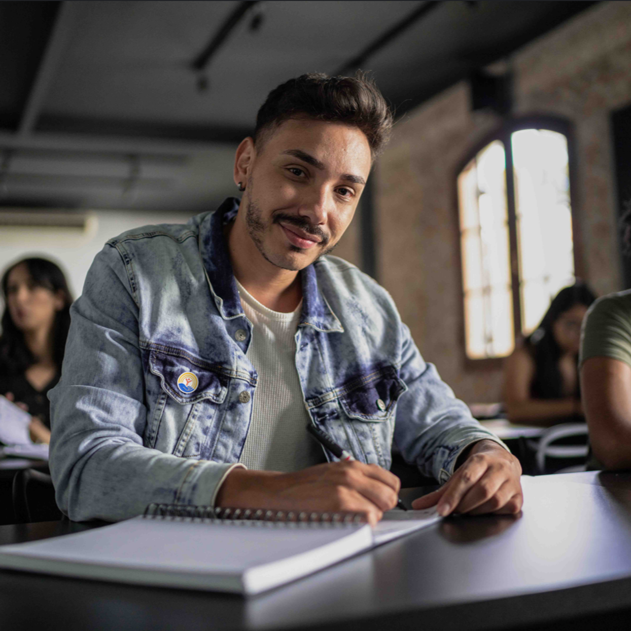 Student sitting at a desk taking notes with a United Way pin on their jean jacket. Student is looking and smiling at camera.