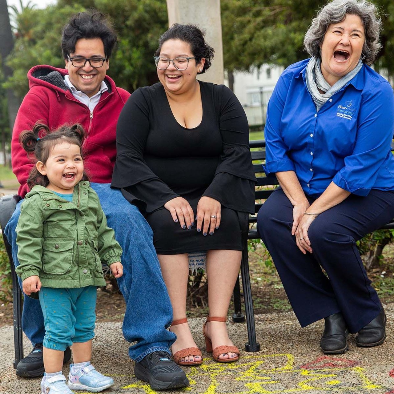 Young parents and a Nurse-Family Partnership nurse are seated on a bench, laughing alongside the smiling toddler who stands in front of them.
